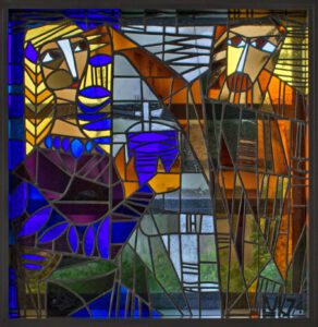 Ein Glasfenster zeigt eine Frau mit einem Ölgefäß und einen Mann, der seine Hand an ihren Kopf hält