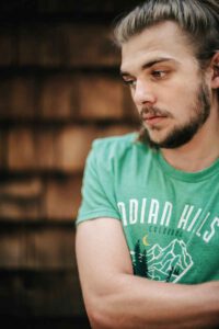 Ein junger Man im grünen T-Shirt und Bart schaut mit gesenktem Blick zu Boden.
