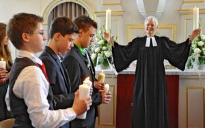 Eine lächelnde Pfarrerin hebt ihre Hände. Im Vordergrund stehen Jungen mit Kerzen