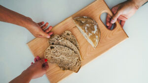 Mehrere Hände halten ein Holzbrett mit aufgeschnittenem Brot und einem Brotstück von oben betrachtet.